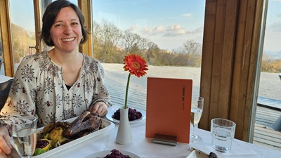 Frau sitzt im Restaurant Oktogon am Himmel, vor ihr das Bio Weidegans Gericht mit Rotkraut, eine orange Blume und die Karte hinter ihr die Aussicht auf Wien