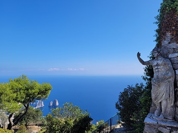 Ein Blick vom Berg aus auf das Meer mit Felsformationen. Links sind Bäume, rechts eine Statue, die ihren Arm hebt.