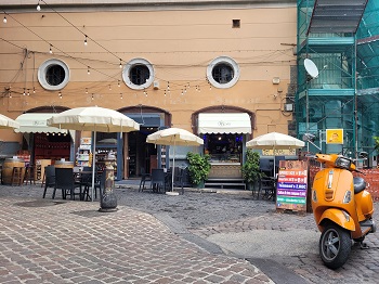 Stühle, Tische und Schirme stehen vor einem Lokal auf der Gasse. Ein oranges Moped steht neben einem Schild, das Getränke ab € 1 anpreist.