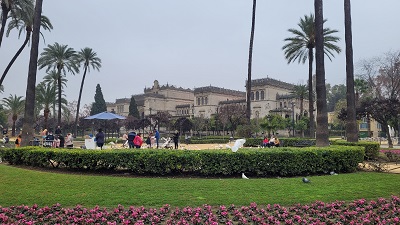 Gepflegter Rasen, gesäumt von Blumen, weiße Tauben fliegen und dahinter liegt ein historisches Gebäude mit für Andalusien typischer Architektur