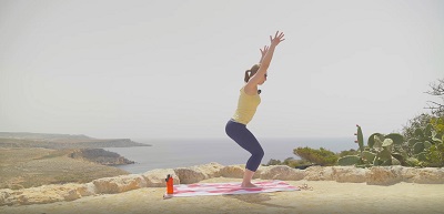 Yogalehrerin zeigt Stuhl Position als Teil des Sonnengrußes B im Vinyasa Yoga auf einem Hügel in Malta, im Hintergrund Klippen und Meer