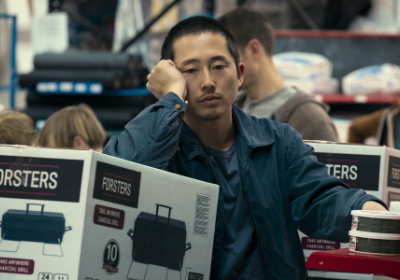 Steven Yeun als Danny in Episode 1 von der Netflix-Serie Beef. Er lehnt auf einem Karton in dem sich ein kleiner Grill befindet. Er sieht unglücklich aus.