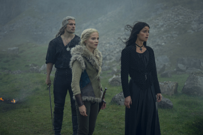Geralt von Rivia mit einem Schwert in der Hand steht neben Yennefer und Ciri auf einer grünen Wiese im Nebel.