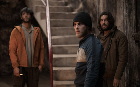 Ein Standbild aus dem Film Bird Box: Barcelona. Drei Männer stehen vor einer Stiege und schauen von rechts nach links.