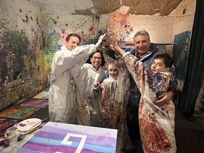 Familie mit bunt bespritzten weißen Anzügen vor einem Bild bei Art Adventure in Graz