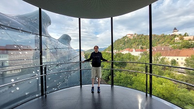Mann steht an der Glasfront im Kunsthaus Graz mit einem prächtigen Ausblick auf den Schlossberg und Uhrturm