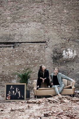Verena Doublier und Sebastian Radon von Wiener Blond sitzen auf einer Couch, neben ihnen ist ein Porträt
