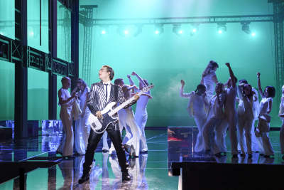Ein Foto während des Musicals ROCK ME AMADEUS. Ein Mann auf der Bühne. Er spielt im Rampenlicht die Gitarre, während hinter ihm das Esemble tanzt.