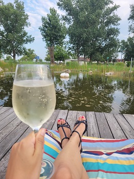 Weinglas mit weißem Spritzer in Damenhand auf Steg mit Teich dahinter