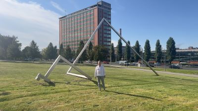 Eine große Wiese mit einer modernen Skulptur. Zwei miteinander verbundene dreidimensionale Dreiecke aus zarten Stahlträgern. Dahinter ein mehrstöckiges schlankes Gebäude.