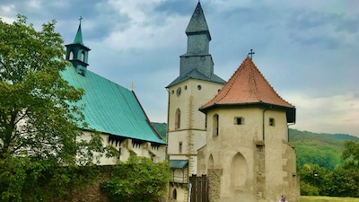 Eine Mauer und ein Wehrturm vor einer gotischen Kirche mit Kirchturm und Dachreiter.