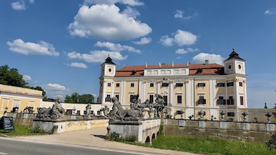 Eine über einen Graben führende Brücke mit barocken Skulpturen. Dahinter ein zweitürmiges Barockschloss mit weißer und crèmegelber Fassade.