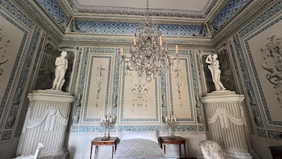 Ein klassizistisch ausgemalter Raum, von der Decke hängt ein Kristallluster, in den Ecken zwei Kamine, darauf jeweils Statuen.