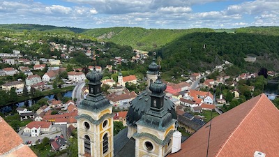Blick auf die Türme einer barocken Kapelle und eine im Tal liegende Ortschaft. Im Hintergrund bewaldete Hügel.