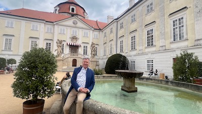 Ein Mann sitzt an der Einfassung eines Brunnens, daneben eine Kübelpflanze. Umgeben von einem barocken Hof, im Hintergrund eine Freitreppe.