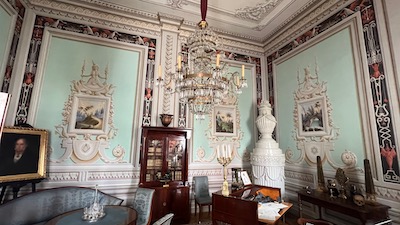 Ein im Stil des Klassizismus eingerichteter Raum mit Kristallluster, weißem Kamin und bemalten Tapeten.