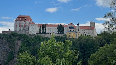 Ein auf einem Felsmassiv stehendes Schloss Vranov. Langgestreckt. Eine Kirche, ein ovaler Bau sind zu sehen. Unterhalb des Schlosses Wald.