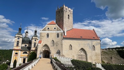 Eine Brücke, die zu einem Burgschloss führt, mit Bauten gotischer Architektur und einer zweitürmigen Barockkapelle. Vorburg zum Schloss Frain