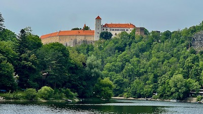 EIn Burganlage, in der Mitte ein viereckiger Turm, thront auf einem steil abfallenden, bewaldeten Hang über einer Wasserfläche.