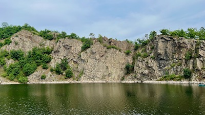 Eine grüne Wasserfläche, am Ufer hoch aufragende Felswände, auf denen Bäume stehen. 