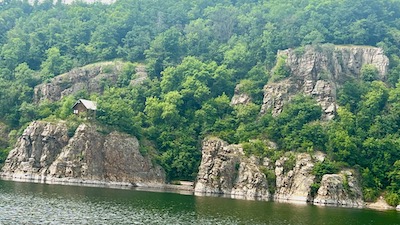 Meterhohe Felsen an einem Seeufer. Darauf steht eine alleinstehende Holzhütte. 