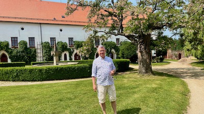 Ein Mann steht in einem begrünten Innenhof. Im Hintergrund ein einstöckiges weißes Gebäude, gedeckt mit rotbraunen Dachziegeln. 