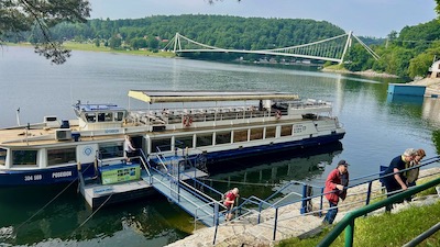 Ein zweideckiges Ausflugsschiff aneinem Landesteg. Einige Passagiere verlassenes über eine Treppe eine Böschung hinauf. Im Hintergrund eine weiße Hängebrücke. 