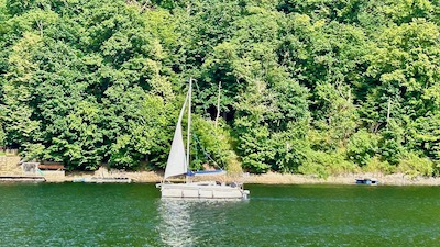 Ein kleine weißes Seegelschiff auf einem See vor einem begrünten Ufer.
