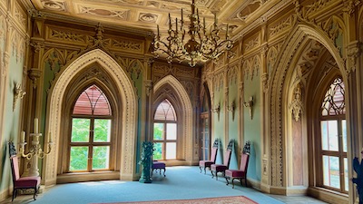 Ein mit Fresken, die den Eindruck von gotischem Masswerk erzeugen sollen. An der Wand Wandleuchten und hohe Lehnsessel. An der Decke ein mehrarmiger Leuchter.