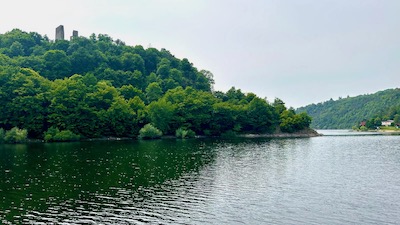 Die Ruine einer mittalterlichen Burgwand ragt aus einem begrünten Hügel am Rand einer Wasserfläche.
