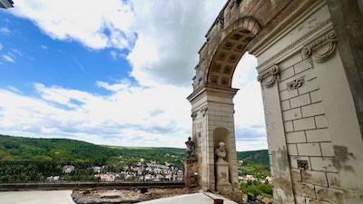 Ein antikisierendes Tor, künstliche Ruine, auf einer Terrasse, mit Blick zu bewaldeten Hügeln.