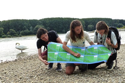 Zwei Helden der Freizeit Redakteur:innen bekommen von Rangerin Kerstin Drabits am Donau-Ufer vor einem Schwan die Nationalpark-Karte erklärt.