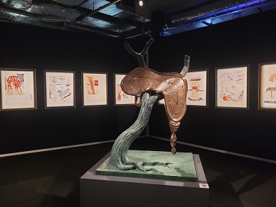 Skulptur einer schmelzenden Uhr auf einem Baum, im Hintergrund gerahmte Bilder auf schwarzer Wand bei der Dalí Ausstellung in Wien