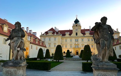 Ein Barockschloss, davor zwei barocke Statuen. In Abendbeleuchtung in der Dämmerung.