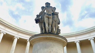 Drei Steinfiguren nackter Frauen vor einem weißen Säulengang und hellblauem bewölkten Himmel.