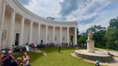 Eine runde Säulenhalle der Nachbildung eines griechischen Tempels. Viele Radfahre legen Rast ein und sitzen auf Stufen. Davor ein Denkmal mit drei unbekleideten Frauen.