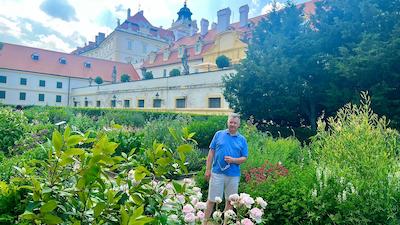 Ein Mann mit blauen Polo steht inmitten von Kräutern und Blumen vor den Seitenflügeln eines Schlosses.