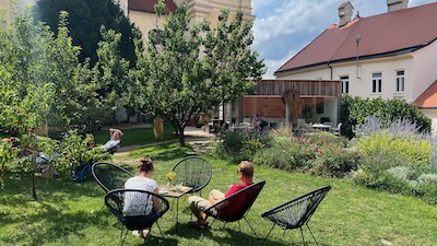 Menschen in bequemen Gartenstühlen auf der grünen Wiese eines Gartencafés neben einer Barockkirche.
