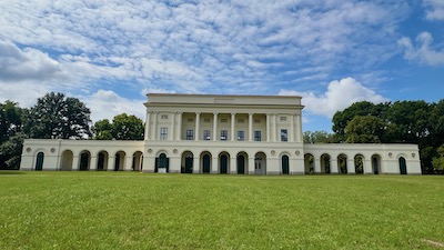Ein langgestreckts Empireschloss mit zwei Seitenflügeln. Davor eine große grüne Wiese. Dahinter blauer, leicht bewölkter Himmel.
