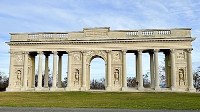 Eine klassizistische, monumental Gloriette auf einer grünen Wiese , dahinter strahlend blauer Himmel.