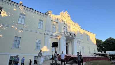 Ein zweistöckiges barockes Schloss. Die weiße Fassade leuchtet in der Abendsonne. Dahinter strahlend blauer Himmel.