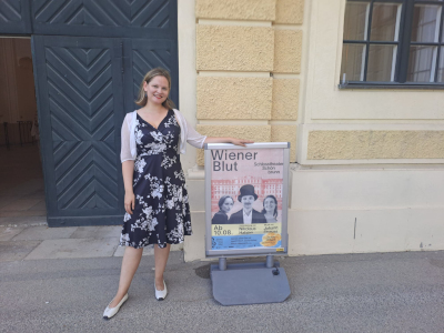 Frau in schwarz-weißem Kleid, lächelnd, vor dem Eingang des Schlosstheaters Schönbrunn, davor das Werbeplakat zur Operette "Wiener Blut"