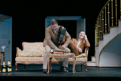 Theaterszene zeigt cremefarbenes Sofa, auf dem ein Mann in braunem Gewand sitzt, auf dem Tisch liegt ein Aktenordner...der Mann legt beinahe seine Füße auf den Ordner, aber ein zweiter Mann (stehend) hebt dessen Füße an, neben dem Sofa: ein Tisch mit Alkoholflaschen, blaue Wand mit weißen Türen (offen) und daneben eine vergoldete Wendeltreppe