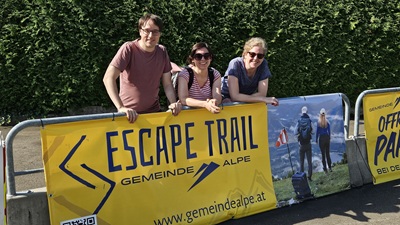 Ein Mann und zwei Frauen mit Sonnenbrille vor einem Plakat des Escape Trails in Mitterbach