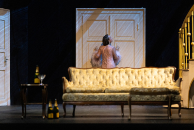 Theaterszene zeit cremefarbenes Sofa, daneben ein Tisch mit Alkoholflaschen, hinter dem Sofa: weiße Türen auf blauer Wand, vor der eine Frau in einem rose Kleid steht, daneben eine vergoldete Wendeltreppe