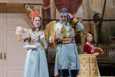 Theaterszene zeigt drei Figuren: In der Mitte ein grauhaariger, bebrillter Mann in Uniform (weiß mit goldenen Epauletten und ein wenig Rot), daneben links eine Frau in einem weißen Kleid mit roter Haarschleife, daneben rechts Frau in rotem Kleid mit Fächer....alle blicken verdutzt drein und stehen vor einer Sitzgarnitur in Epaulettenform....im Hintergrund: eine weiße Tür, an der Bühnendecke: eine Spiegelfläche, die den Saal des Schlosstheaters Schönbrunn teilweise reflektiert