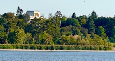 Ein griechischer Tempel, versteckt hinter Bäumen auf einem sanften Hügel vor einem Teich.