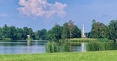 Blick über eine Wiese und einen Schlossteich zu einem Aqädukt und einem Minarett. Strahlend blauer Himmel.