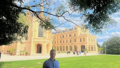 Mann in hellblauem Hemd unter Baumästen vor grüner Wiese. Dahinter ein in heller Crémefarbe gestrichenes Schloss im Stil der Neo-Tudorgotik.