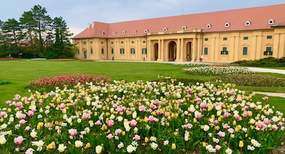Blick über ein großes Blumenbeet und eine grüne Wiese zu der gelben Fassade eines einstöckigen langgezogenen Barockbaus.